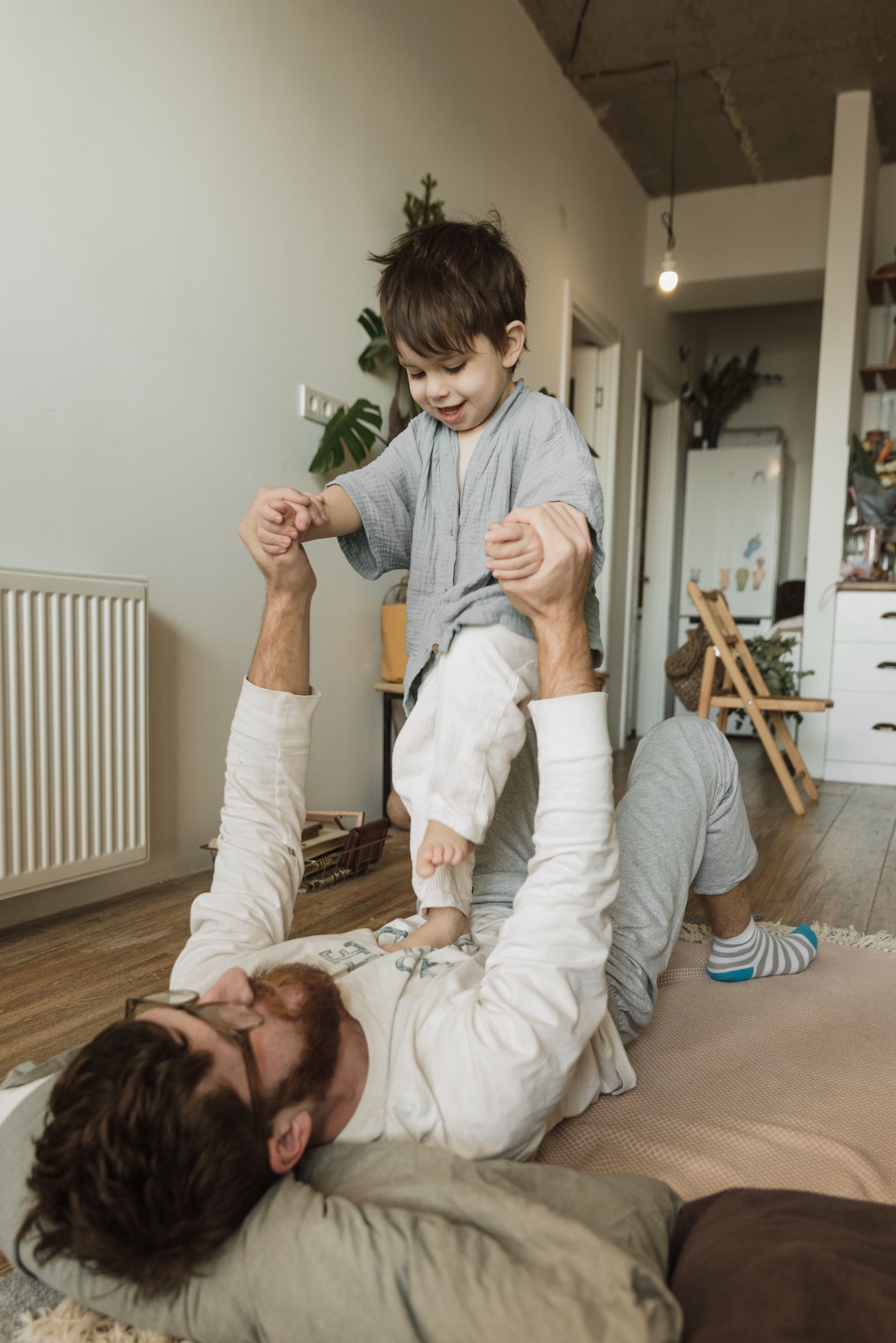 A father lies on a living-room floor holding the hands of a young child standing above him.