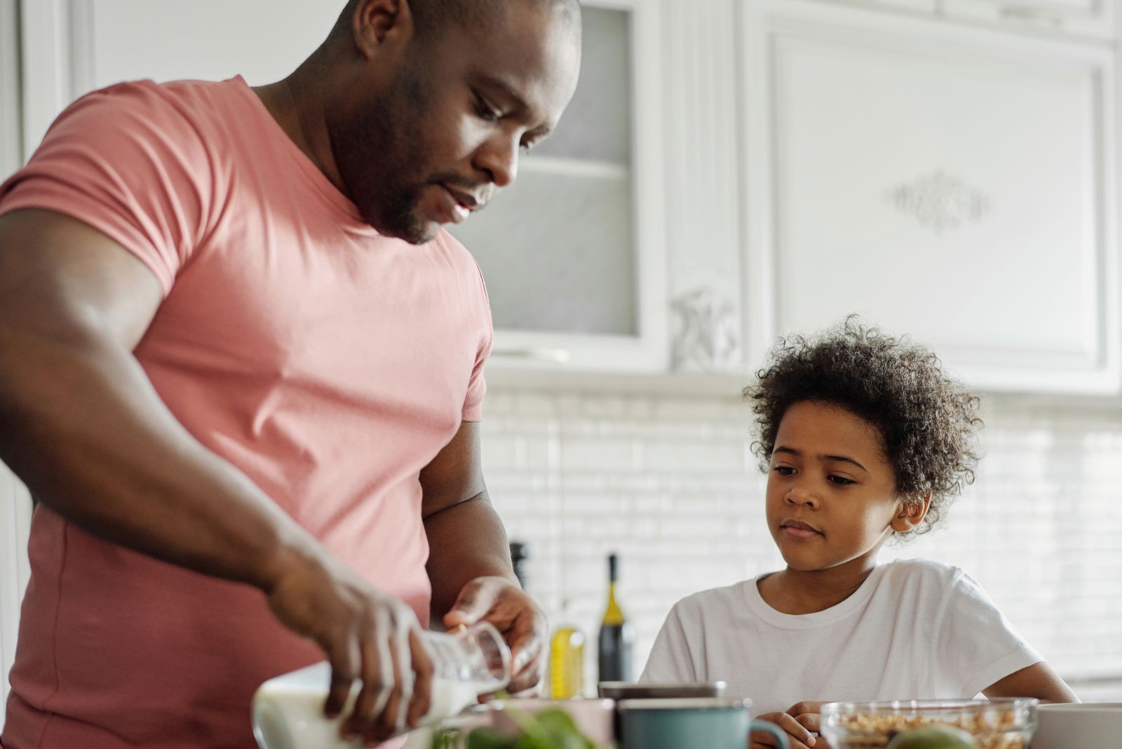 A father pours milk into a bowl while his son watches beside him in a kitchen.