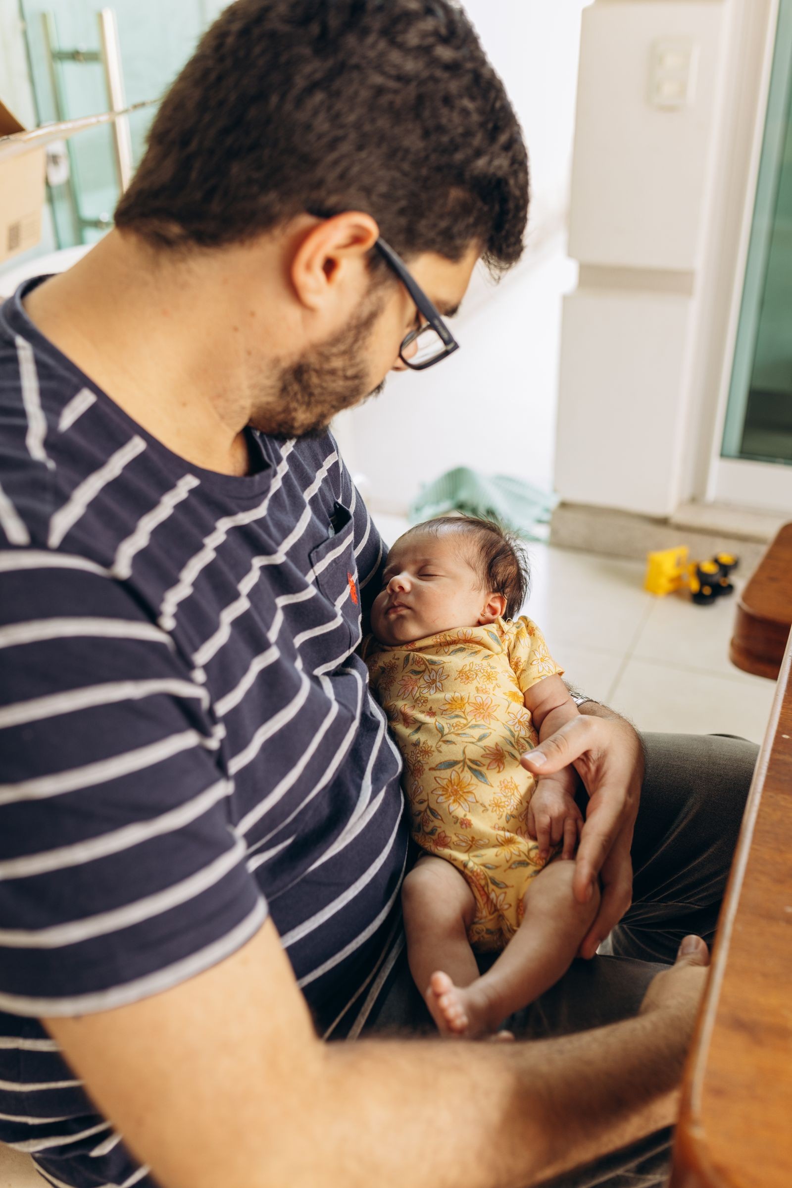 A father seated indoors cradles a sleeping infant in his arms.