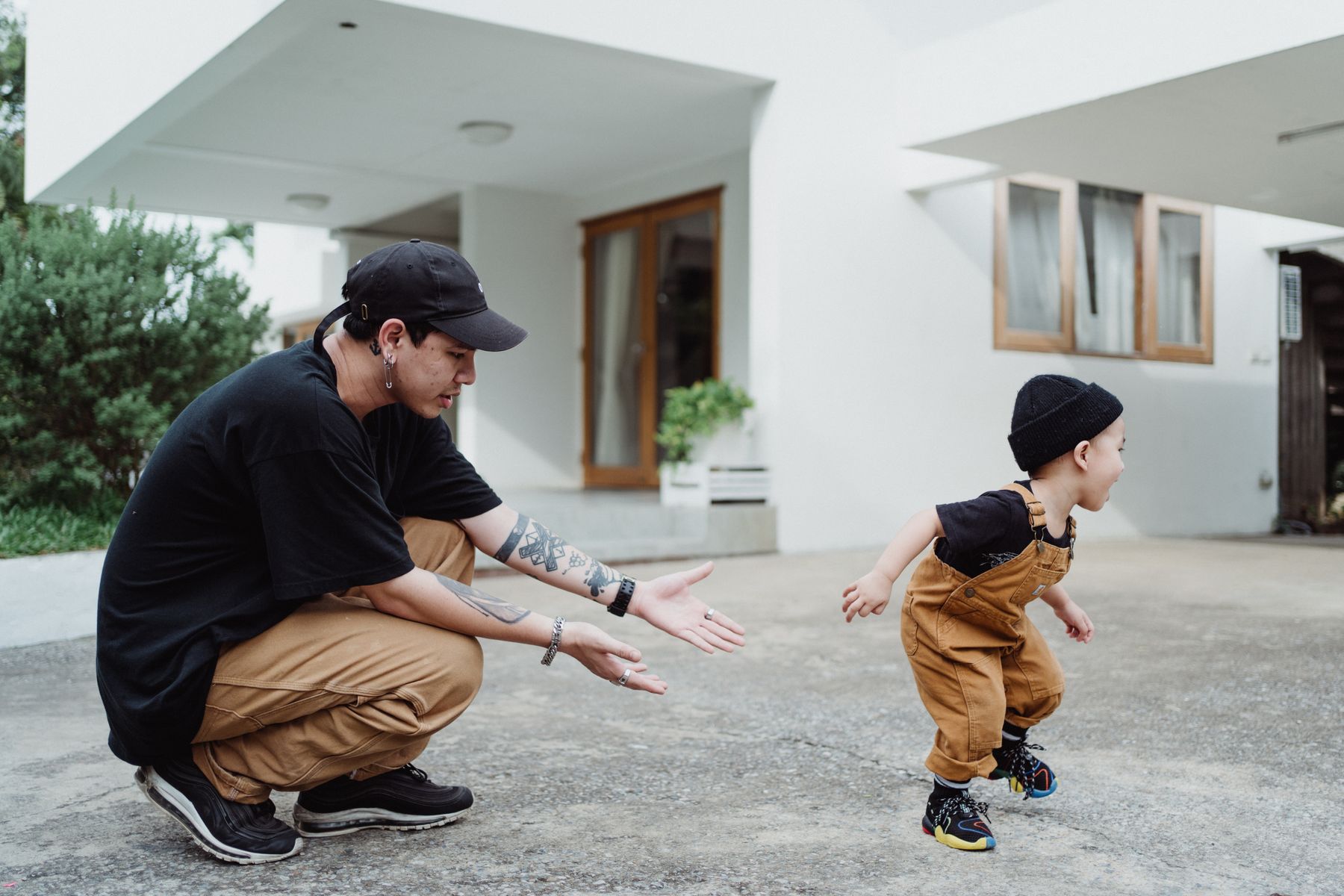 A father crouches with his arms outstretched beside a toddler playing outside a home.
